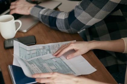 Close-up of hands organizing tax forms on a desk with a calculator, laptop, and notebook.