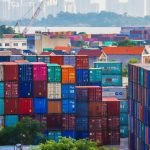 Vibrant stacked cargo containers at a bustling port with urban skyline in background.