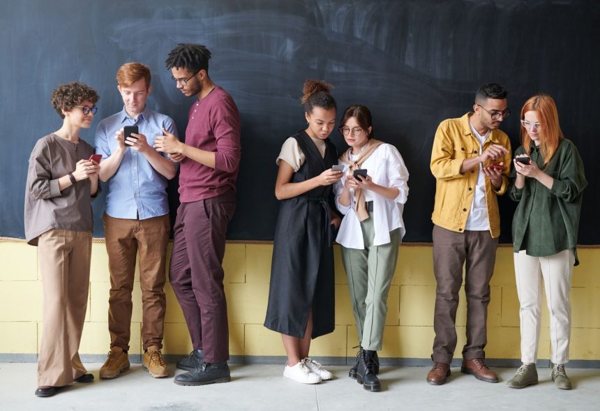 Young diverse adults using smartphones while standing against a blackboard, showcasing technology and connection.